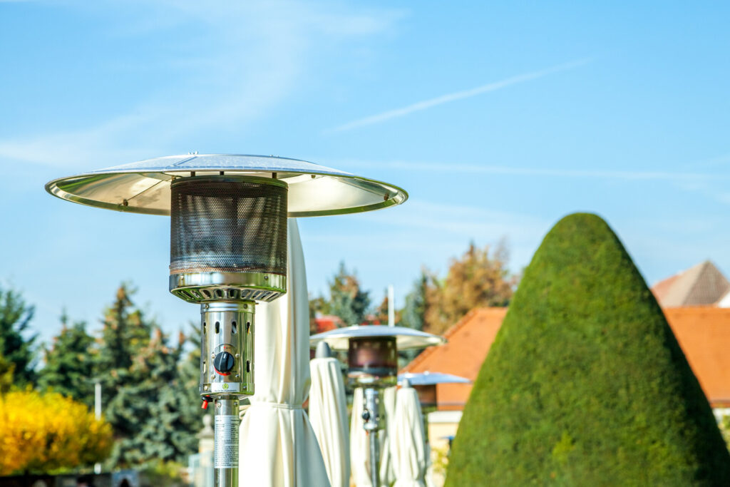 Heizpilz im Freien: Effiziente Heizlösung für Terrassen, betrieben mit Gas oder Petroleum, an einem sonnigen Tag in einem Garten mit grüner Hecke und blauen Himmel im Hintergrund.